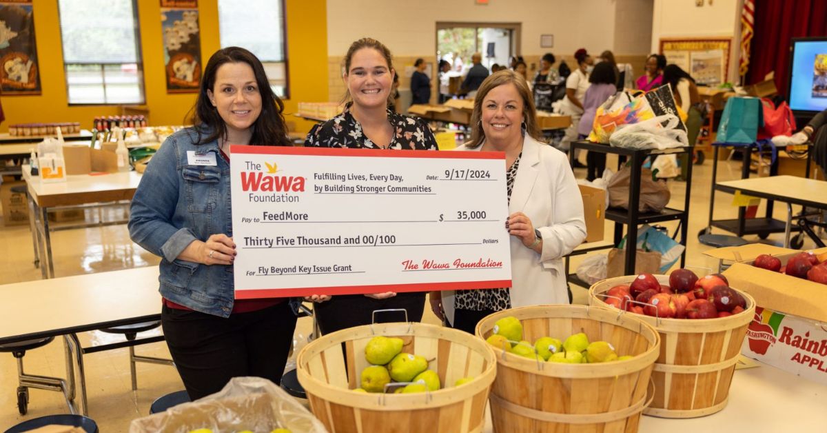 Three smiling women standing behind baskets of fruit holding a big check for a generous grant from the Wawa Foundation at a Feed More distribution.