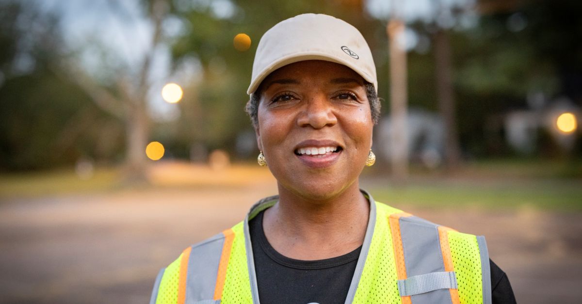 A smiling woman wearing a hat with a safety vest that is dedicated too running a food distribution center. Proving that if you are looking for kindness, look local!