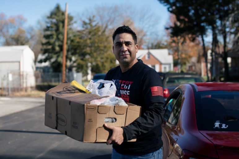A man carrying a box of food donations outside, standing next to a parked car at an agency partner.
