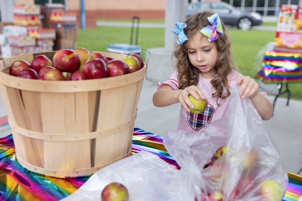 A young girl with colorful hair bows packing apples into a bag at a Feed More children's program food distribution event.
