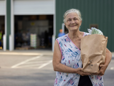An elderly woman smiling while holding a bag of groceries received from Feed More, standing in front of a green warehouse.