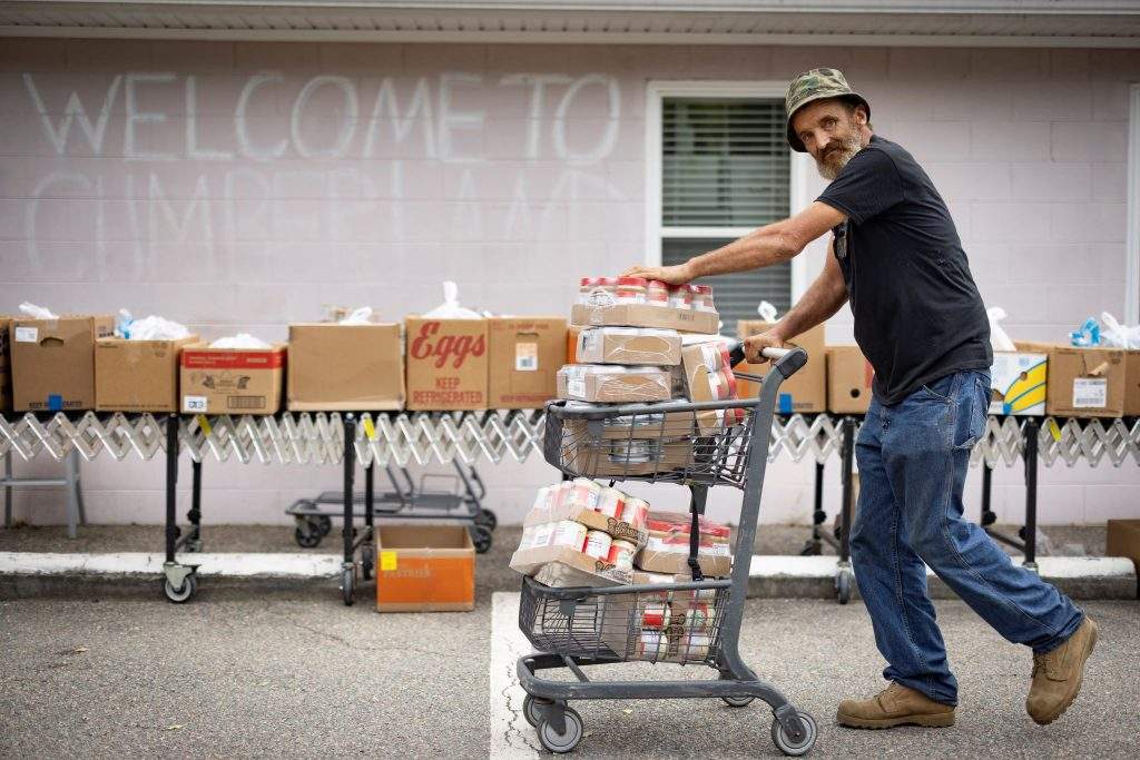 A man pushing a cart filled with food donations at a Feed More distribution event, standing in front of a 'Welcome to Cumberland' sign.