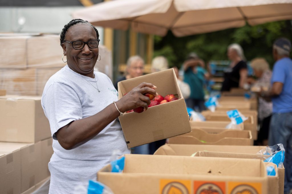 A woman smiling while holding a box of apples at a Feed More food distribution event, with others in the background.