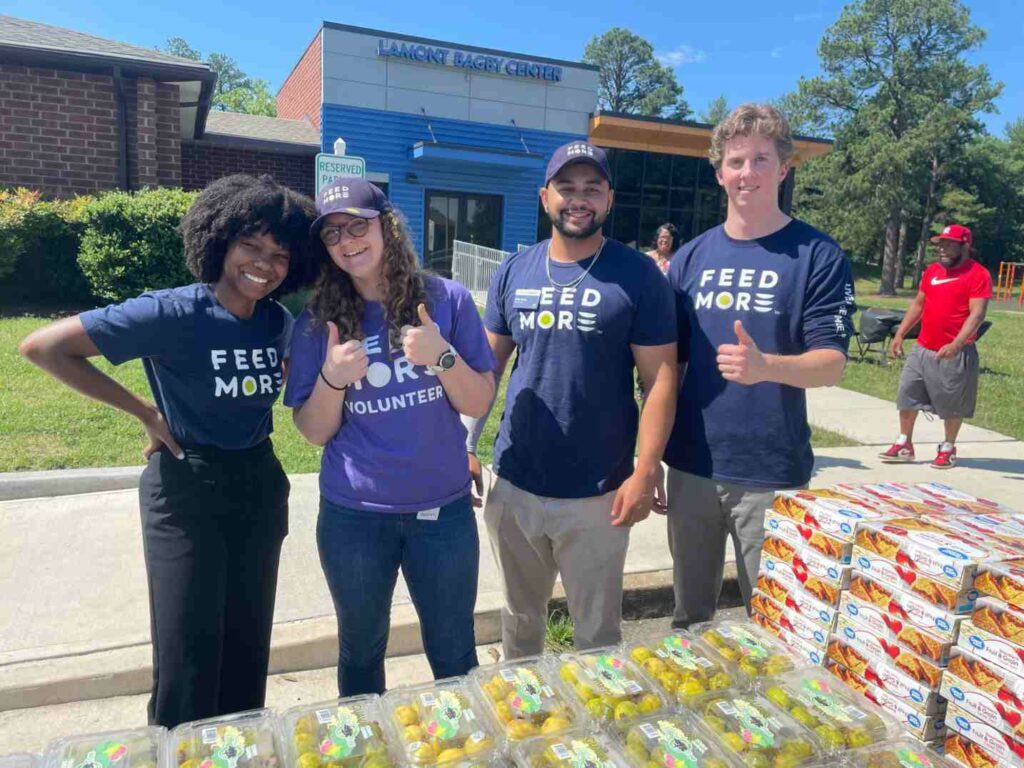 A group of Feed More volunteers standing outside, smiling and giving thumbs up during a food distribution event.